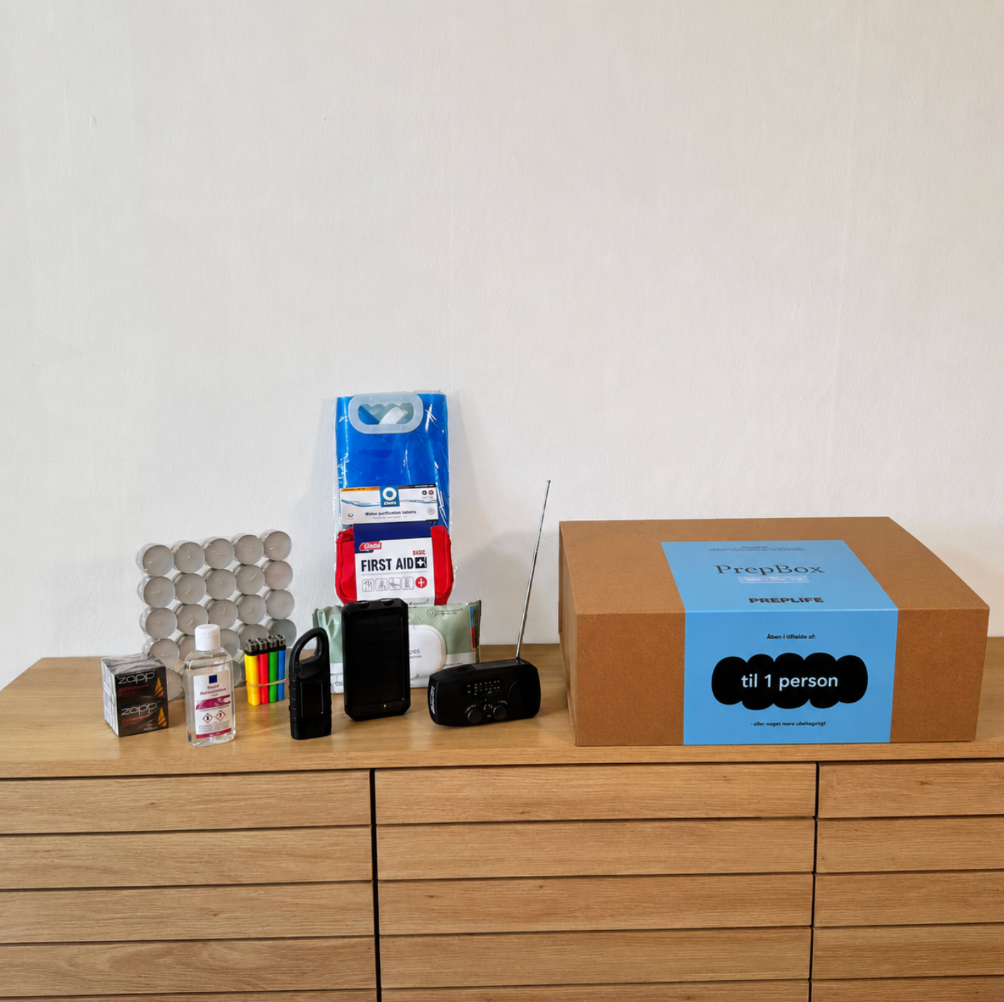 PrepBox One opstillet på en kommode med beredskabsudstyr som fyrfadslys, håndsprit, lommelygter, powerbanks, førstehjælpssæt, nødradio, nødtæpper og foldbare vanddunke ved siden af lukket PrepBox-kasse.