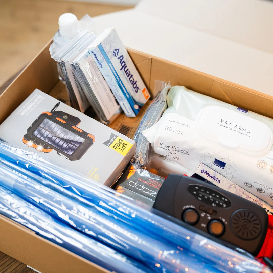 Åben PrepBox med beredskabsudstyr som nødradio, vandrensningstabletter, vådservietter, nødtæpper og foldbare vanddunke samlet i en papkasse.