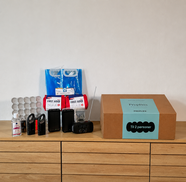 PrepBox Double opstillet på en kommode med beredskabsudstyr som fyrfadslys, håndsprit, lommelygter, powerbanks, førstehjælpssæt, nødradio, nødtæpper og foldbare vanddunke ved siden af lukket PrepBox-kasse.