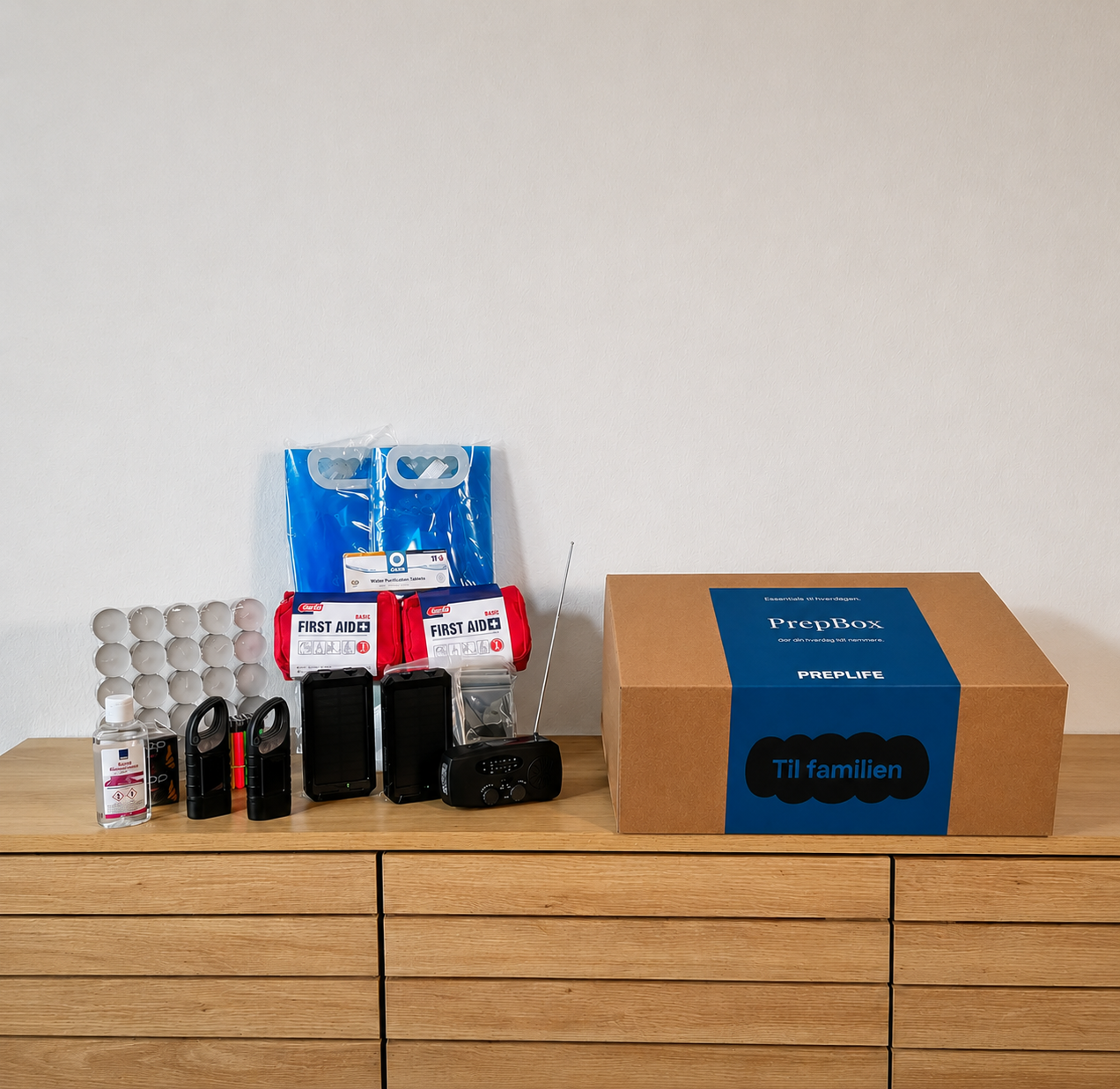 PrepBox Family opstillet på en kommode med beredskabsudstyr som fyrfadslys, håndsprit, lommelygter, powerbanks, førstehjælpssæt, nødradio, nødtæpper og foldbare vanddunke ved siden af lukket PrepBox-kasse.